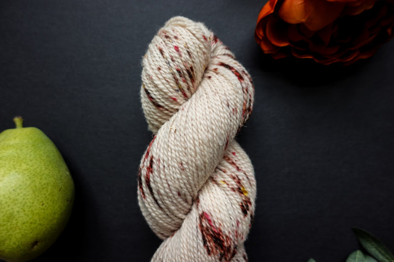 A naturally dyed skein of white yarn with burgundy speckles lays on a black surface. An orange-red flower and a pear sit next to it.
