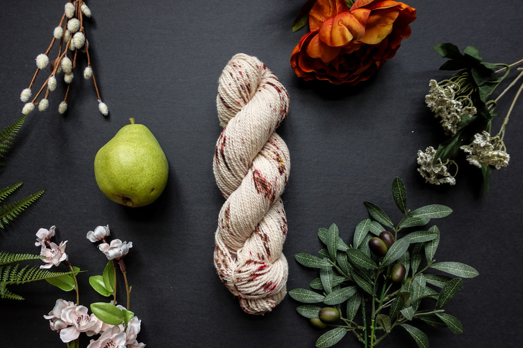 A skein of white DK weight yarn with burgundy speckles lies on a black surface. It's surrounded by flowers, branches, an orange rose, and a pear.