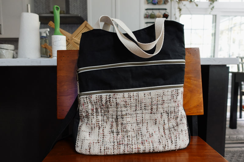 A black canvas and white speckled fabric yarn project bag sits on a chair. There are two front zippered pockets, two woven white handles, and a black canvas sling strap.