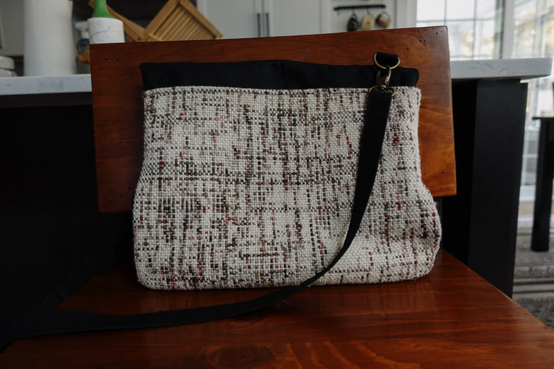 Pictured from the bag, and black canvas and white, speckled, handwoven fabric sits on a chair. It has a long black canvas strap that attaches with bronze lobster clasps.