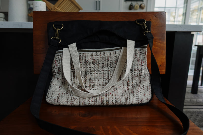 A black canvas project bag sits on a chair. It has a front zippered pocket made of a speckled white, handwoven fabric. There are two white handles, and a long black canvas strap connected with two bronze lobster clasps.