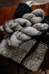 Two speckled skeins of white yarn lay on top of a knitting project bag. The bag is made of black canvas and a speckled white handwoven fabric that matches the yarn. 