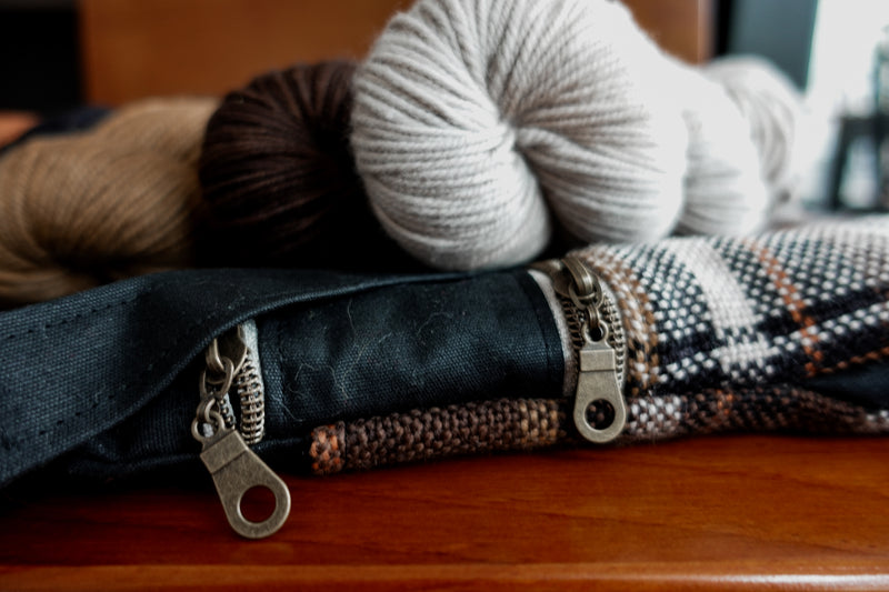 A black canvas and orange and white flannel yarn project bag sits on a chair with three yarn skeins. The camera is close up to the two zipper toggle closures.