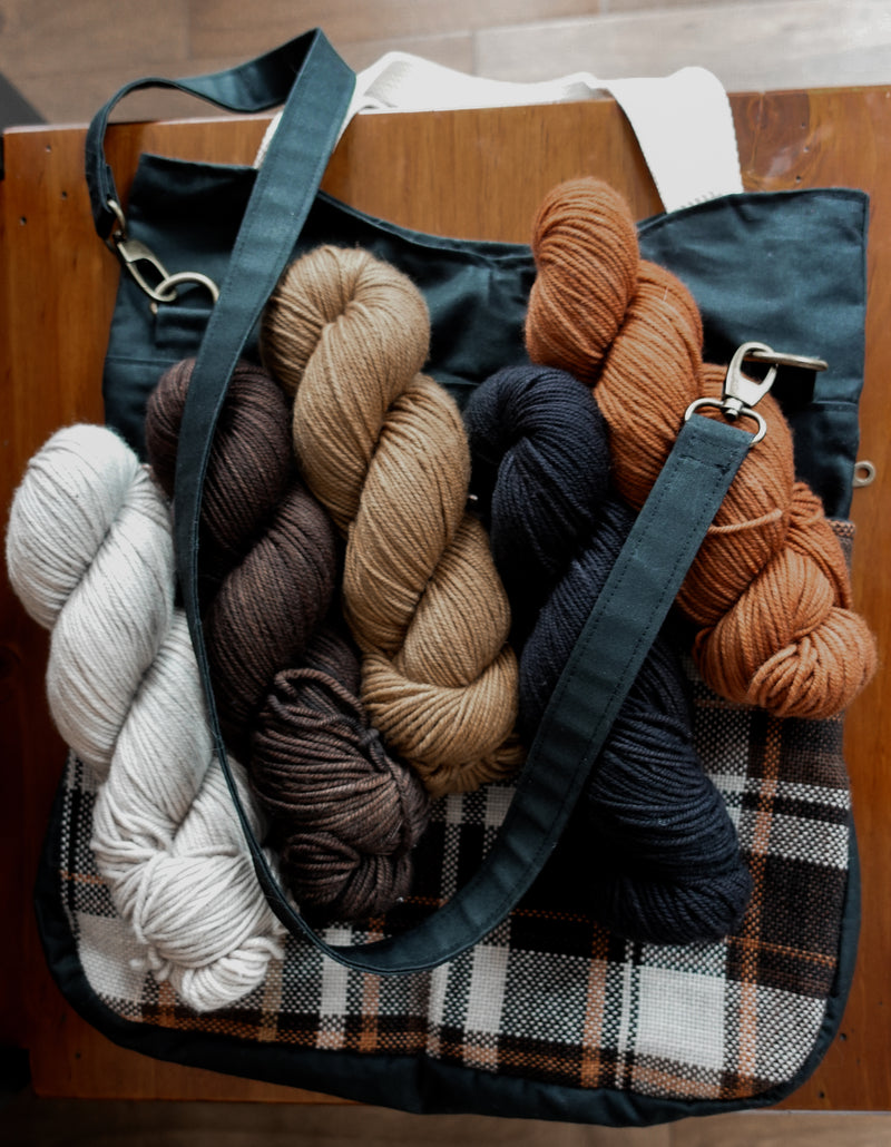 A handwoven black and flannel project bag lays on a chair. Five skeins of yarn in complimenting colors sit on top, showing that they would fit comfortably in the bag.
