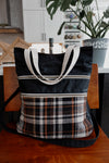 A knitting project bag, made of black canvase and orange and white flannel, sits on a chair. It has two front pockets, white handles, and a long black sling strap.