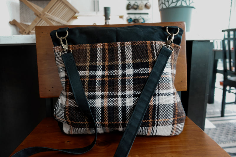 A black canvas and orange flannel knitting project bag sits on a chair. It has a long black sling strap, attached to the bag with two bronze lobster clasps.