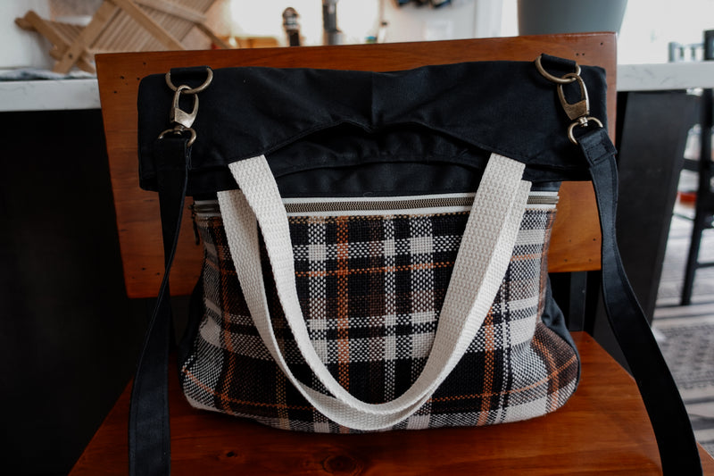 A black canvas project bag sits on a chair. It has a handwoven flannel zipper pouch on the front, white tote handles, and a black sling strap.