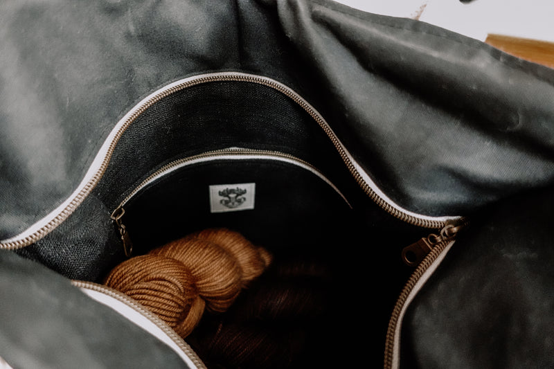 The zippered top of a handmade knitting project bag is open to show the black linen lining inside. There is a zippered interior pocket, and a small version of the Forest Lane Fiber Co. logo sewn inside. A skein of yarn has been placed inside, showing the large amount of room.