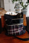 Seen from the front, a black canvas and purple flannel project bag sits on the chair. It has two zippered front pockets, with a skein of hand dyed yarn in each one. 
