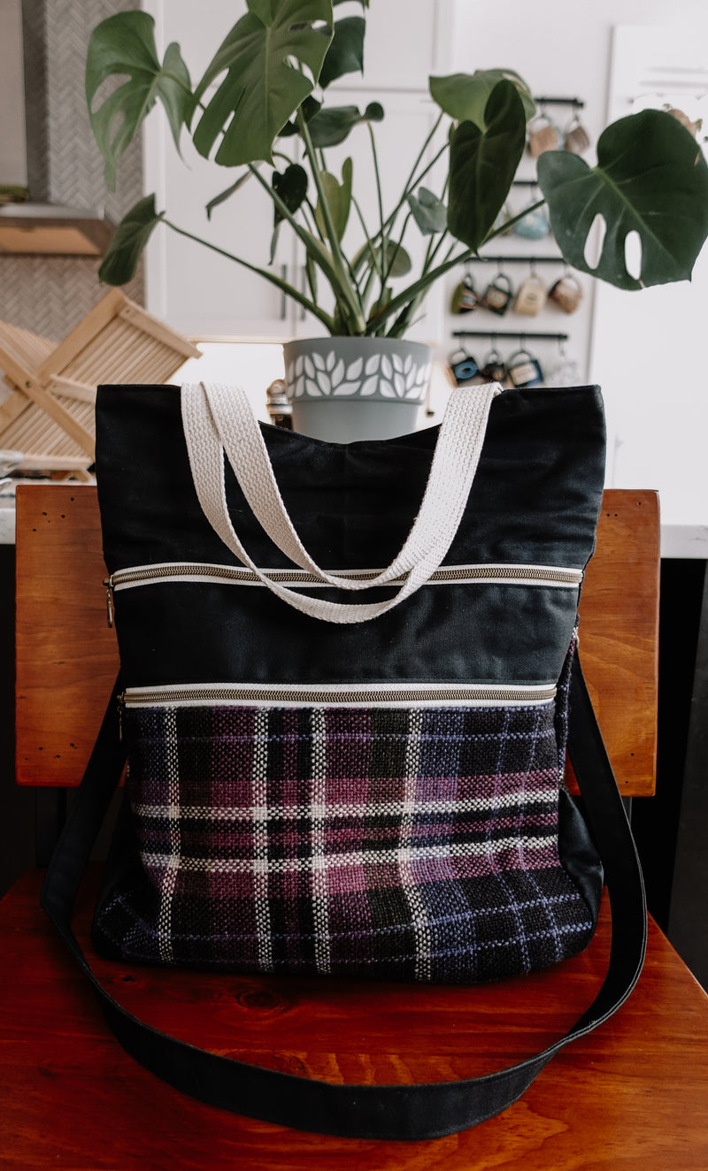 Seen from the front, a black canvas and purple flannel project bag sits on a chair. It has two white woven handles, two front zippered pockets, and a long black canvas sling strap.