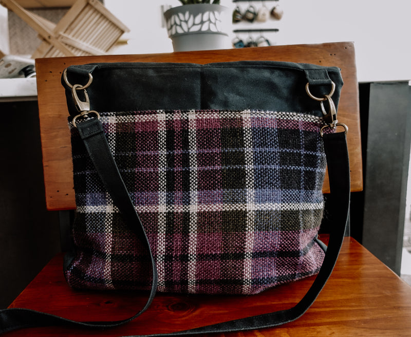 Seen from behind, a black canvas project bag with a handwoven purple flannel bottom half sits on a chair. It has a long black canvas strap that attaches with two bronze lobster clasps.