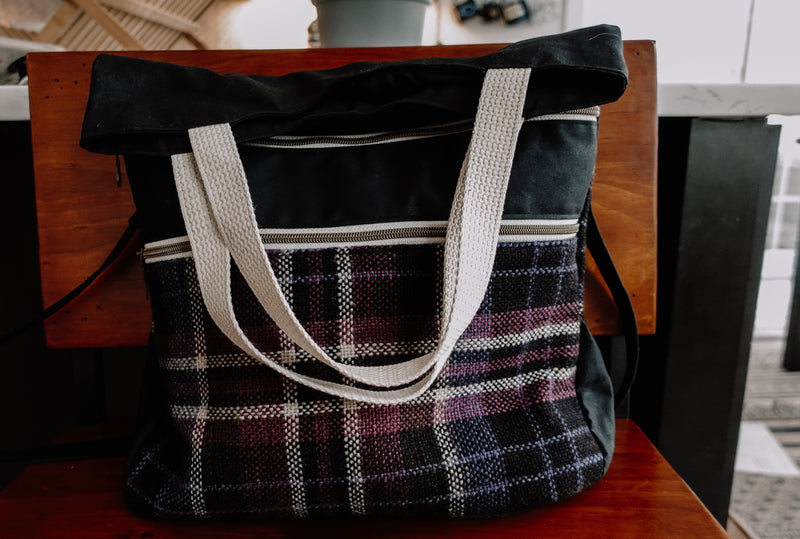 A knitting project bag tote sits on a chair. The front has two zippered pockets with a purple, blue, and white handwoven flannel. The bag has white woven handles and a black sling strap.