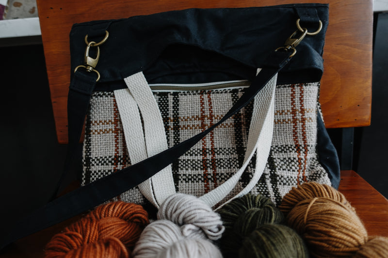 A knitting project bag sits on a chair. It's made of black canvas, and orange and white handwoven flannel. It has two white handles and a black sling strap. Four skeins of yarn sit in front of the bag.