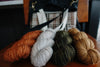 The camera focuses on four skeins of yarn sit in front of a knitting project bag. The knitting project is made of black canvas, and orange and white handwoven flannel. It has two white handles and a black sling strap.