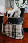 Seen from an angle, a black canvas and white and orange flannel project bag sits on a chair. The two front pockets are unzipped, and each have a skein of yarn in them. 