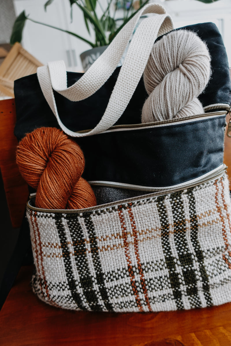 Seen from the front, and orange and white handwoven flannel and black canvas project bag sits on a chair. The two front zippered pockets are open, and both have a skein of yarn in them. 