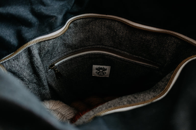 The top pocket of a black canvas knitting project bag is open, showing the inside. It's lined with a herringbone cotton, and has a zippered inside pouch. Two skeins of yarn can be seen at the bottom, showing the large amount of room. 