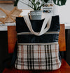 A black canvas yarn project bag with a orange and white flannel pocket, sits on a chair. It has two front zippered pockets, two white woven handles, and a black sling strap.
