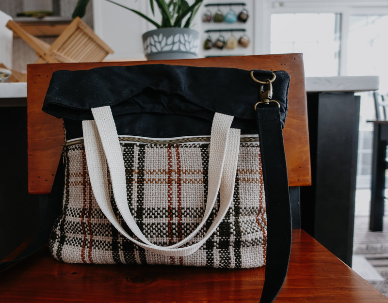 A black canvas knitting project bag sits on the chair. The front pocket is made of handwoven white, orange, and black flannel fabric. The bag has two white handles and a long black canvas sling strap.