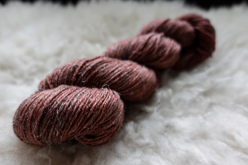 A natural skein of yarn has been hand dyed brick red. It lays on a sheepskin and is seen from the side.