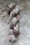 Seen from above, a hand dyed skein of white yarn with burgundy speckles lays on a sheepskin.