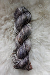 Seen from above, a variegated skein of brown, grey, and white hand dyed yarn lays on a sheepskin.