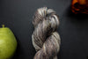 Seen close up, a brown, grey, and white skein of hand dyed, variegated yarn lays next to an orange-red flower and a pear.