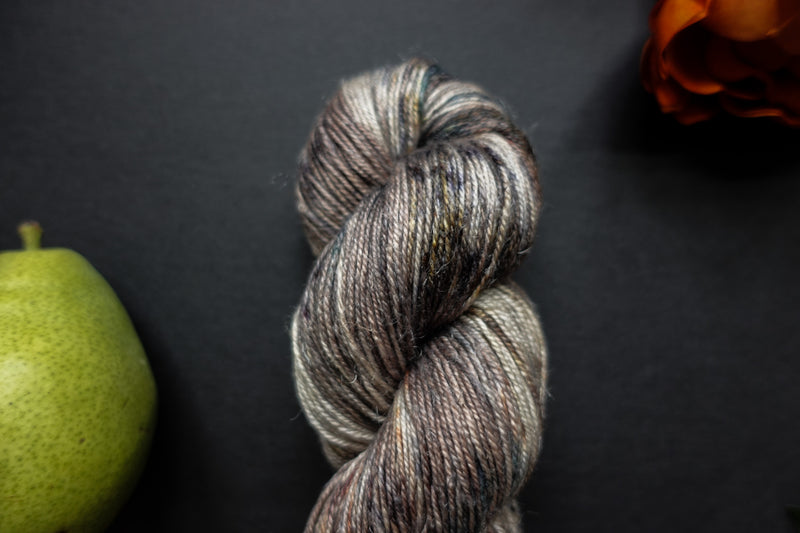 Seen close up, a brown, grey, and white skein of hand dyed, variegated yarn lays next to an orange-red flower and a pear.