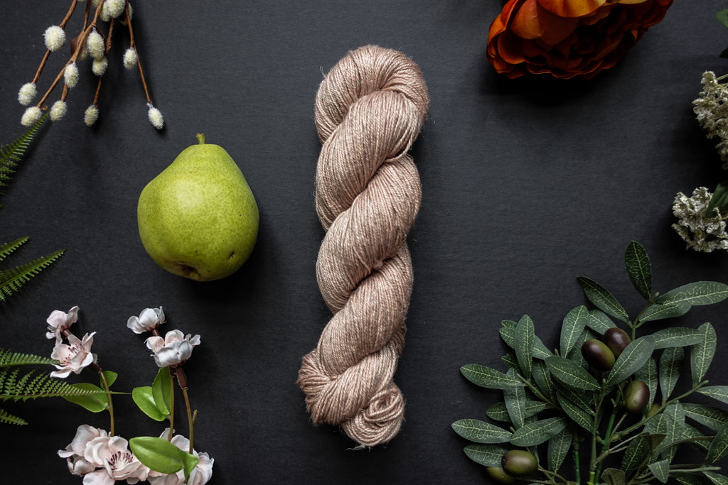A skein of light beige sport weight yarn lies on a black surface. It's surrounded by flowers, branches, an orange rose, and a pear.