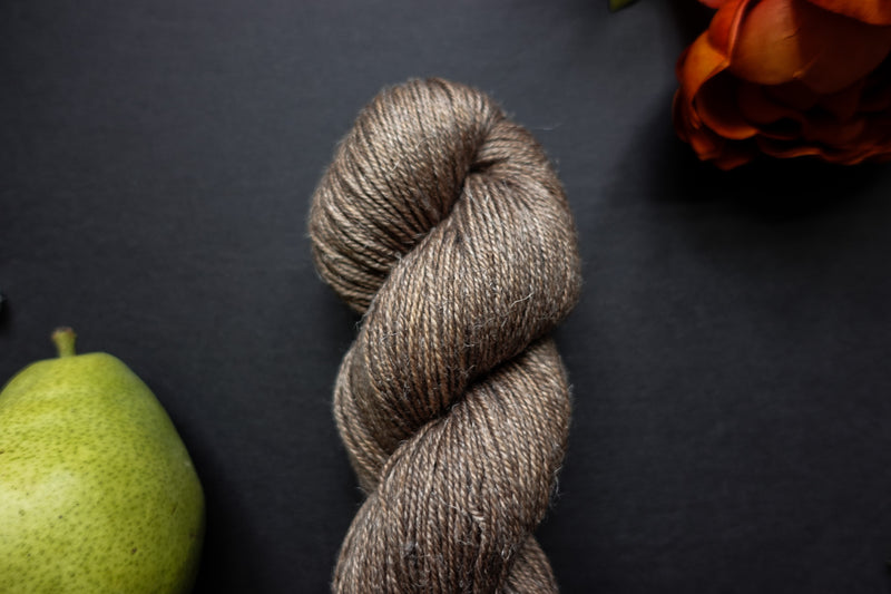 Seen close up, a naturally dyed skein of light brown yarn lays on a black surface next to an orange-red flower and a pear.