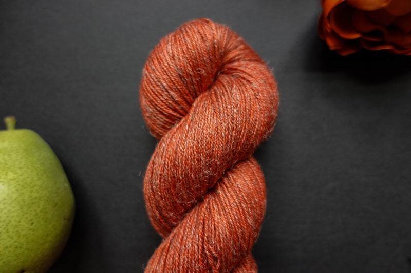 Seen close up, an orange-red skein of hand dyed yarn lays on a black surface next to a matching flower and a pear.