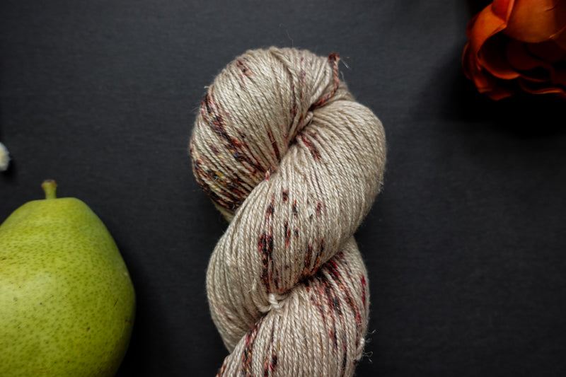 Seen close up, a hand dyed skein of white yarn with burgundy speckles lays on a black surface next to an orange-red flower and a pear.