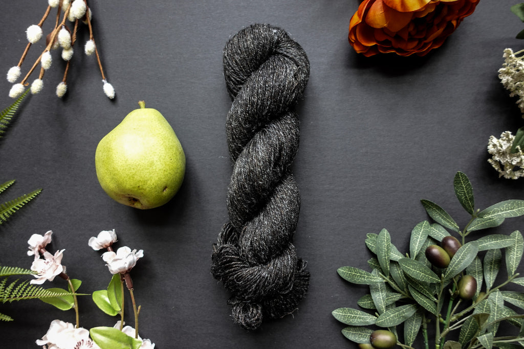 A dark, muddy green, almost black, skein of sport weight yarn lies on a black surface. It's surrounded by flowers, branches, a poppy, and a pear.