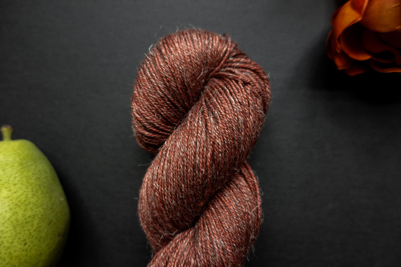 Seen close up, a brick red skein of hand dyed yarn lays on a black surface next to an orange-red flower and a pear.