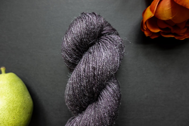 Seen close up, a deep purple skein of hand dyed yarn lays on a black surface next to an orange-red flower and a pear.