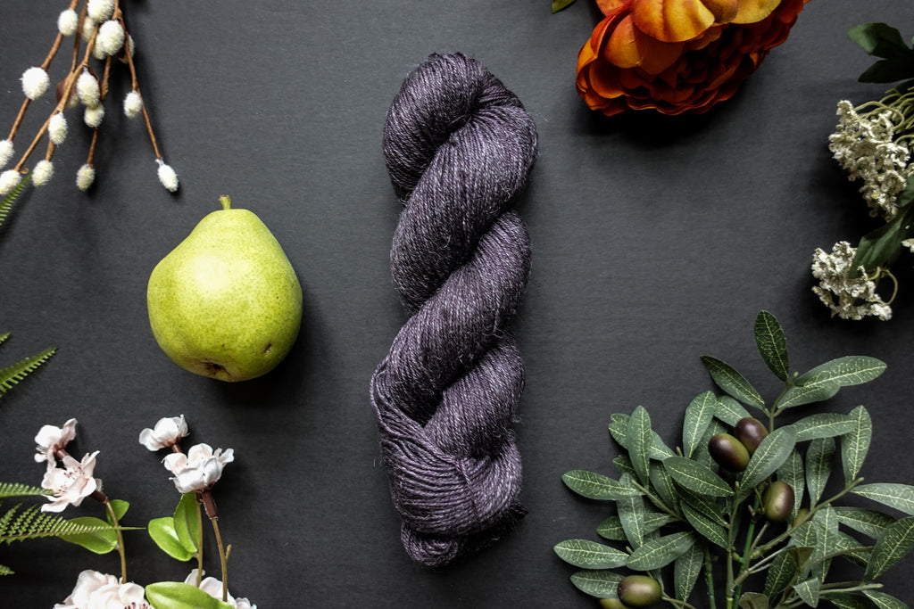 A skein of deep purple sock weight yarn lies on a black surface. It's surrounded by flowers, branches, an orange rose, and a pear.