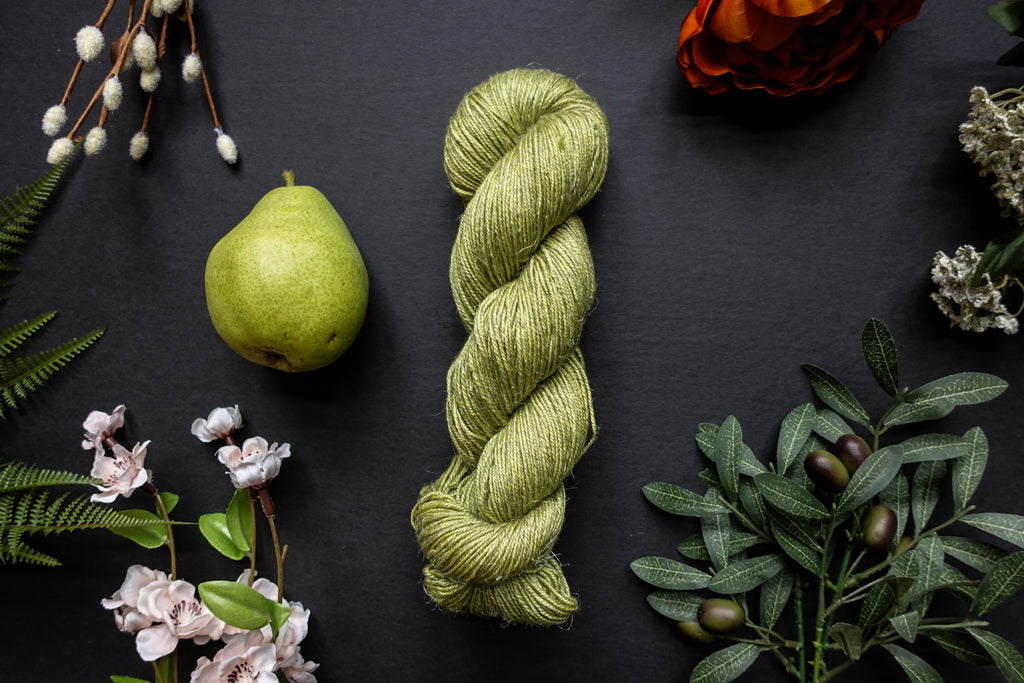 A bright green skein of sport weight yarn lies on a black surface. It's surrounded by flowers, branches, an orange rose, and a pear.