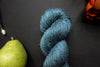 Seen close up, an indigo blue skein of hand dyed yarn lays on a black surface next to an orange-red flower and a pear.