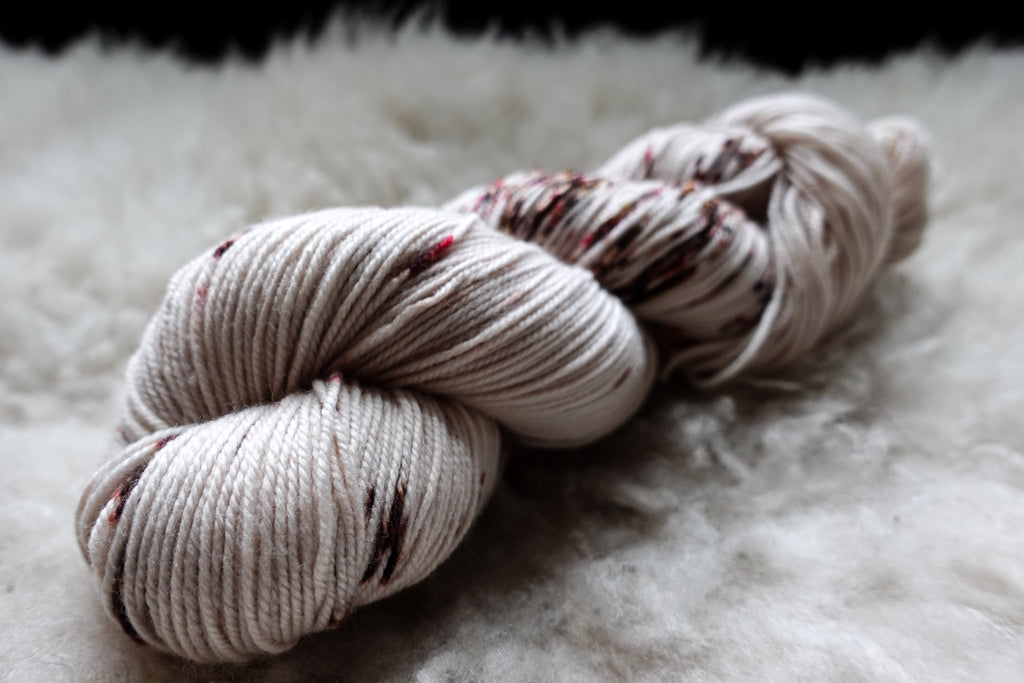 A hand dyed skein of white yarn with burgundy speckles lays on a sheepskin and is seen from the side.