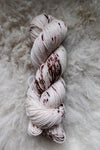 Seen from above, a hand dyed skein of white yarn with burgundy speckles lays on a sheepskin.