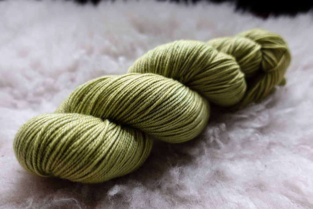 A skein of natural yarn has been hand dyed a bright green. It lays on a sheepskin and is seen from the side.
