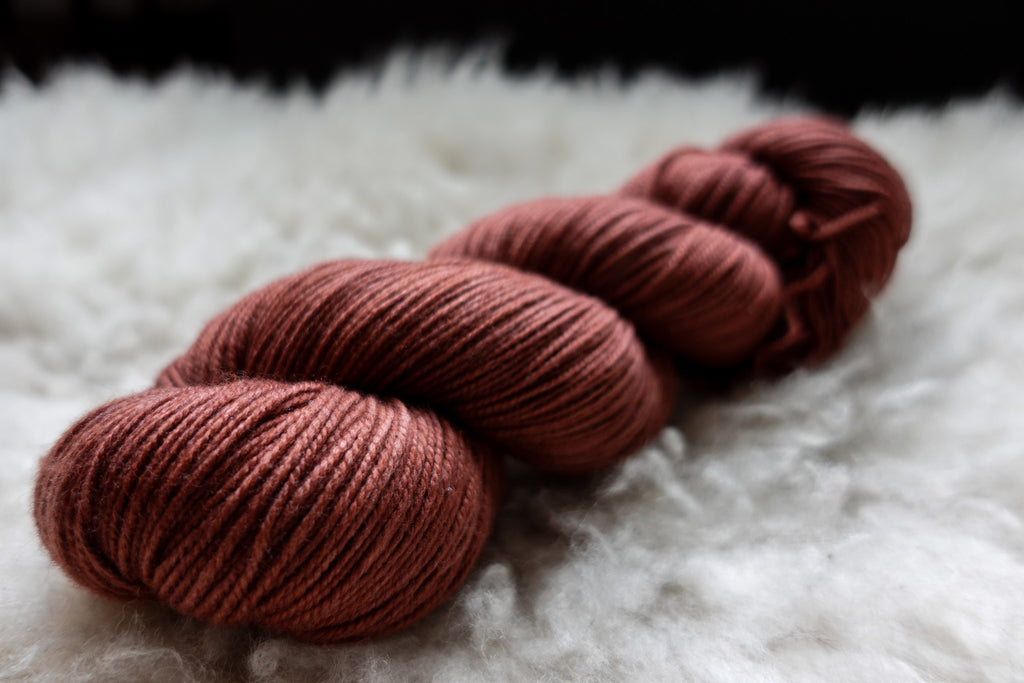 A natural skein of yarn has been hand dyed brick red. It lays on a sheepskin and is seen from the side.