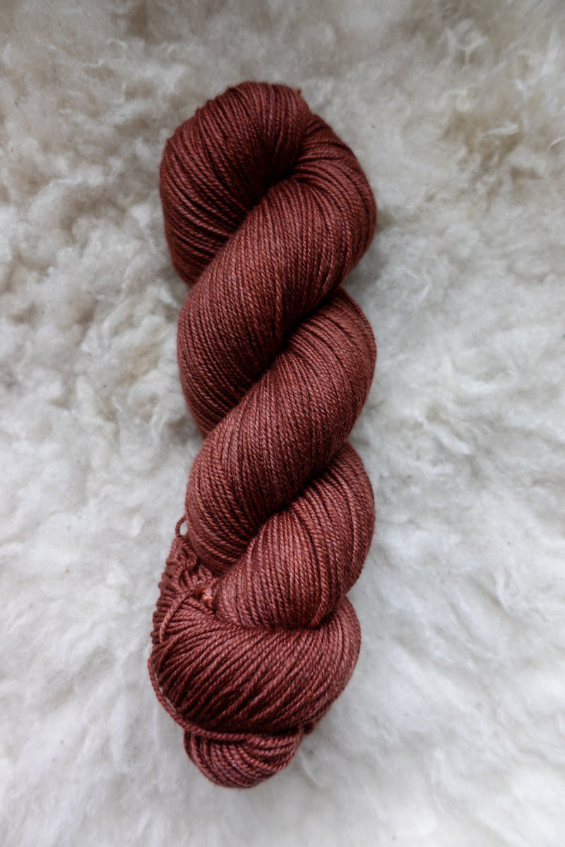 Seen from above, a brick red skein of hand dyed yarn lays on a sheepskin.