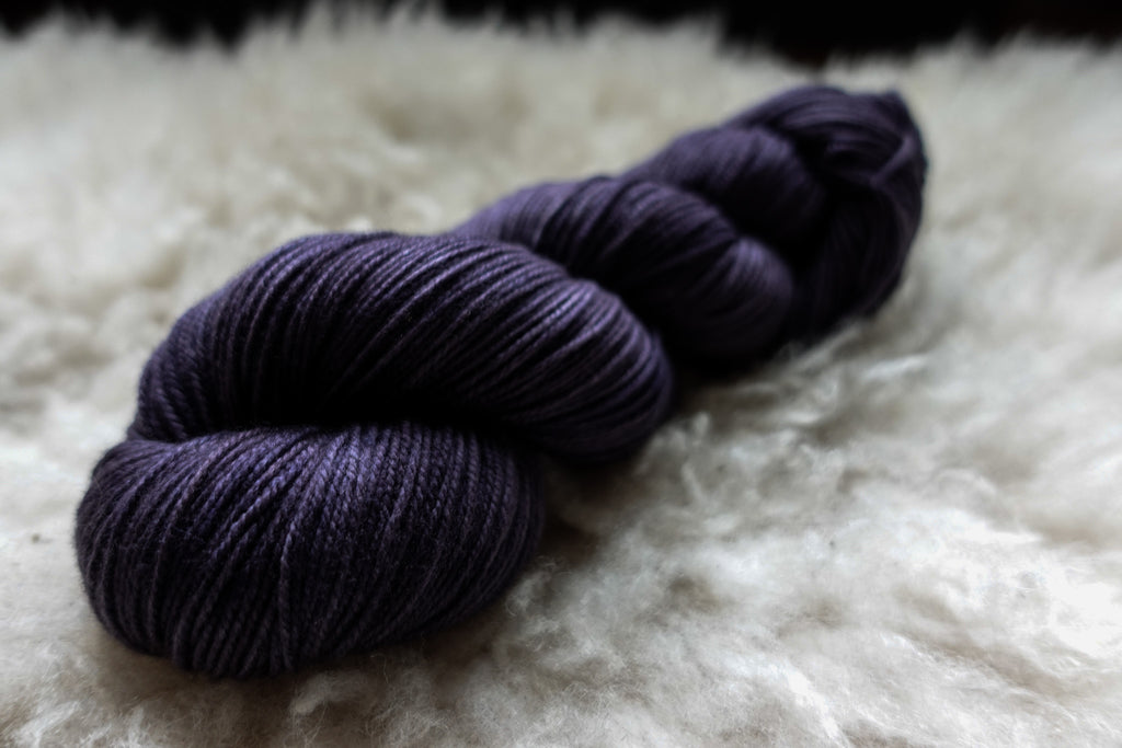 A natural skein of yarn has been hand dyed a deep purple. It lays on a sheepskin and is seen from the side.