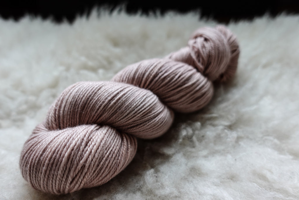 A natural skein of yarn has been hand dyed a light pink-beige. It lays on a sheepskin and is seen from the side.