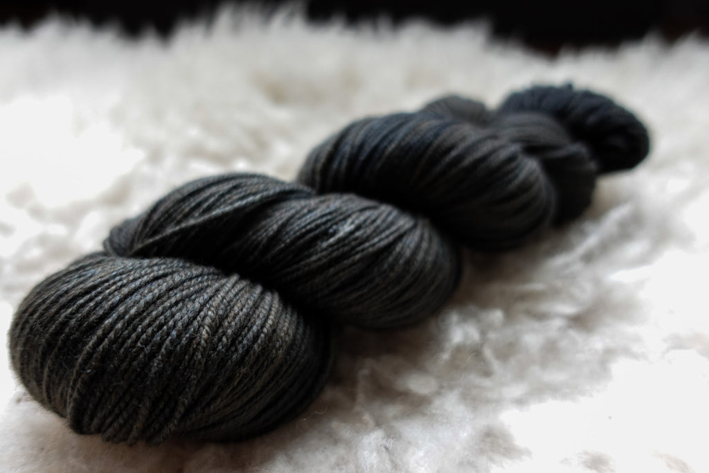 A dark, almost black, skein of natural yarn has been hand dyed. It lays on a sheepskin and is seen from the side.