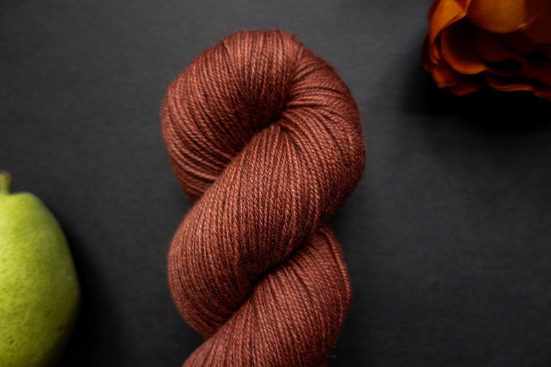 Seen close up, a brick red skein of yarn lays on a black surface next to an orange-red flower and a pear.