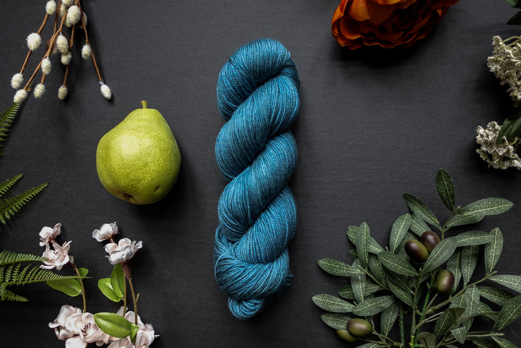A skein of blue sock weight yarn lies on a black surface. It's surrounded by flowers, branches, an orange rose, and a pear.