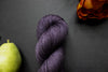 Seen close up, a hand dyed skein of deep purple yarn lays on a black surface next to an orange-red flower and a pear.