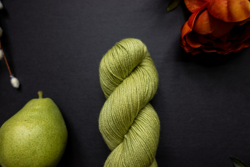 A bright green skein of naturally dyed yarn lays on a black surface next to an orange-red flower and a pear.